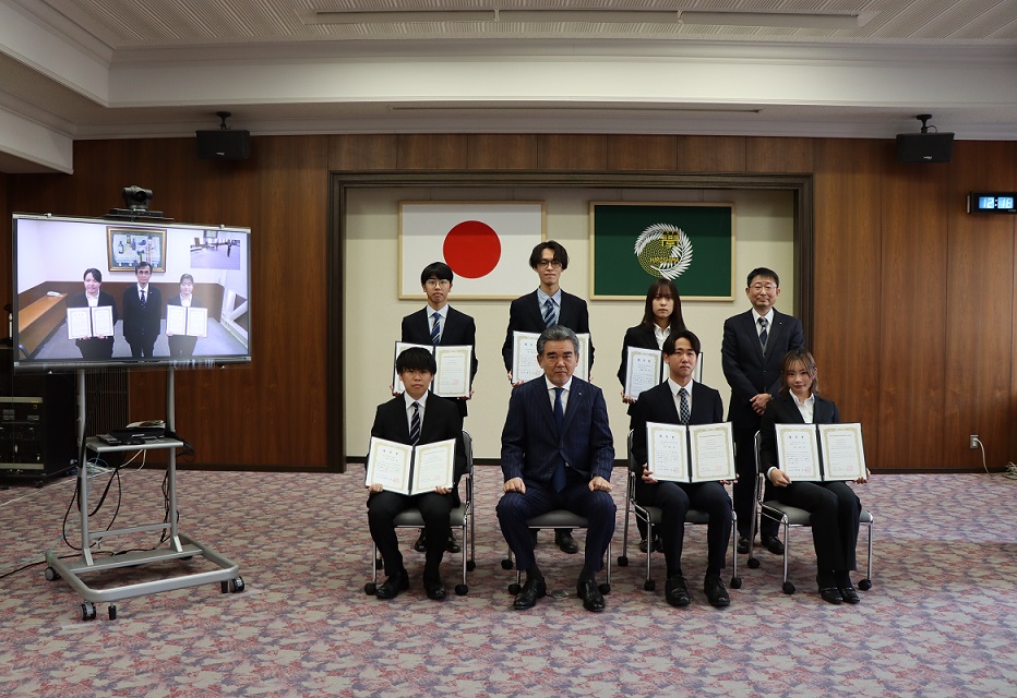 記念撮影(東広島キャンパスの受賞者6人、越智学長、川野平和教育部門長、テレビ画面に映っているのは霞キャンパスの受賞者2人、小澤副学長)