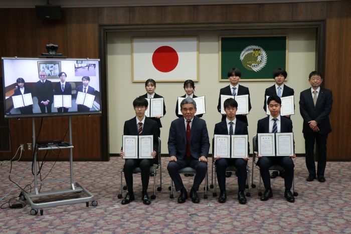 記念撮影(東広島キャンパスの受賞者7人、越智学長、川野平和教育部門長、テレビ画面に映っているのは霞キャンパスの受賞者3人、熊本副学長)